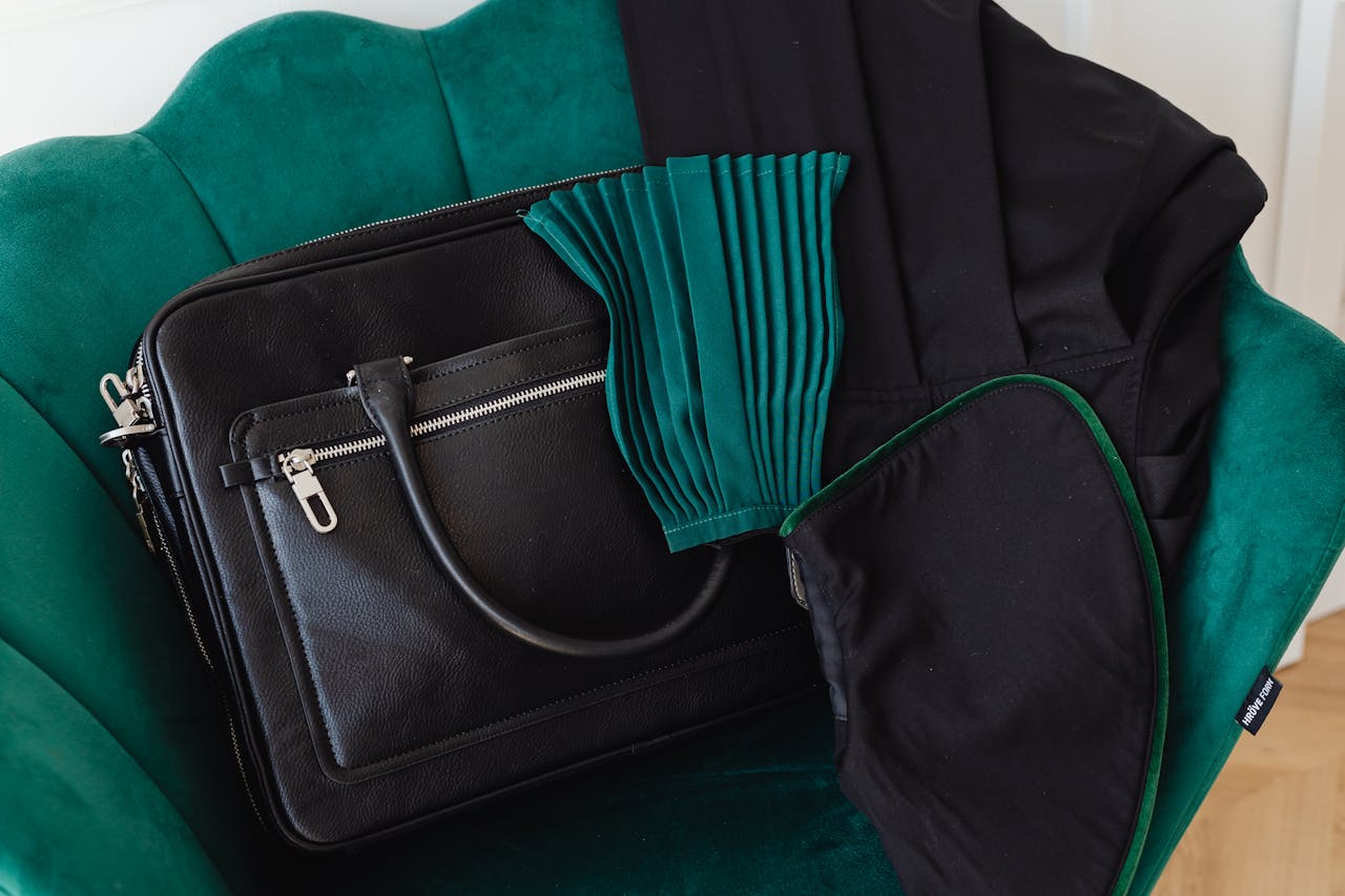 Stylish black leather handbag placed on a luxurious green velvet accent chair with clothing.